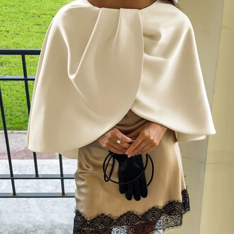 Person wearing a beige cape over a dress with black lace details, holding black gloves.