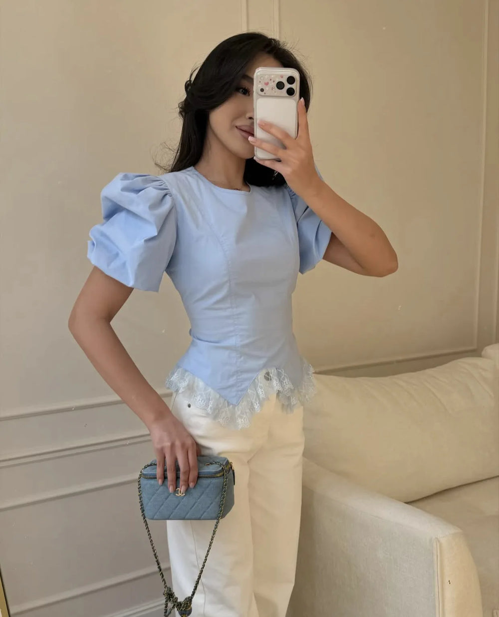Woman taking a mirror selfie wearing a light blue top with ruffled sleeves and white pants, holding a small blue handbag.