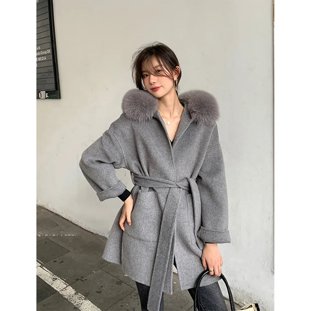 Woman wearing a gray coat with fur hood standing against a plain wall.