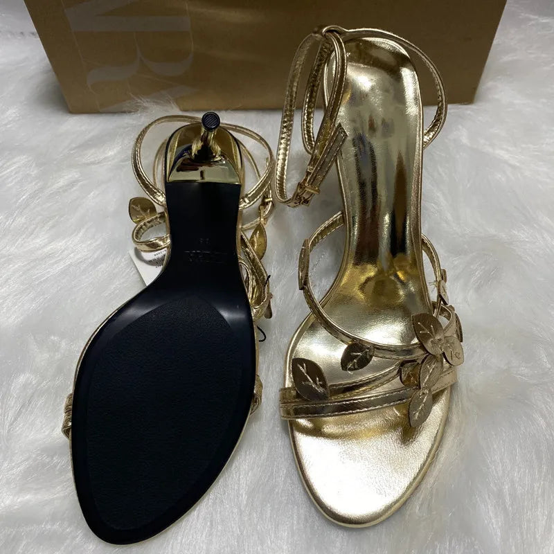 Gold high-heeled sandals with a black shoe tree on a white surface.