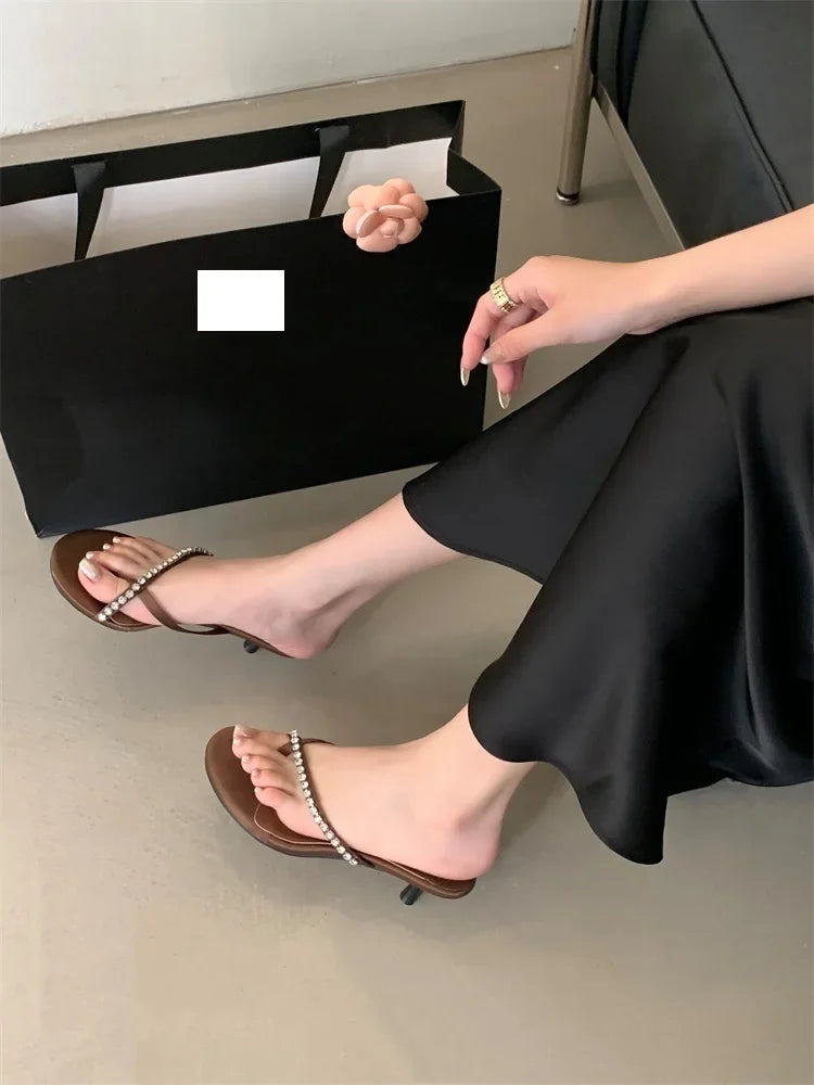 Person wearing brown high-heeled sandals with pearl details, sitting on a black stool.