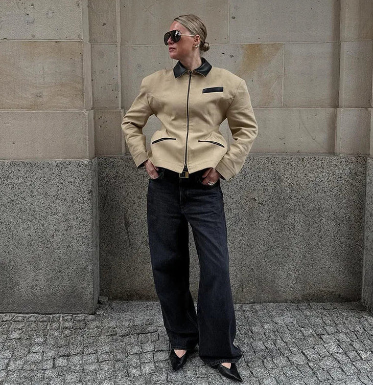 Person wearing a beige jacket with black accents against a stone wall.