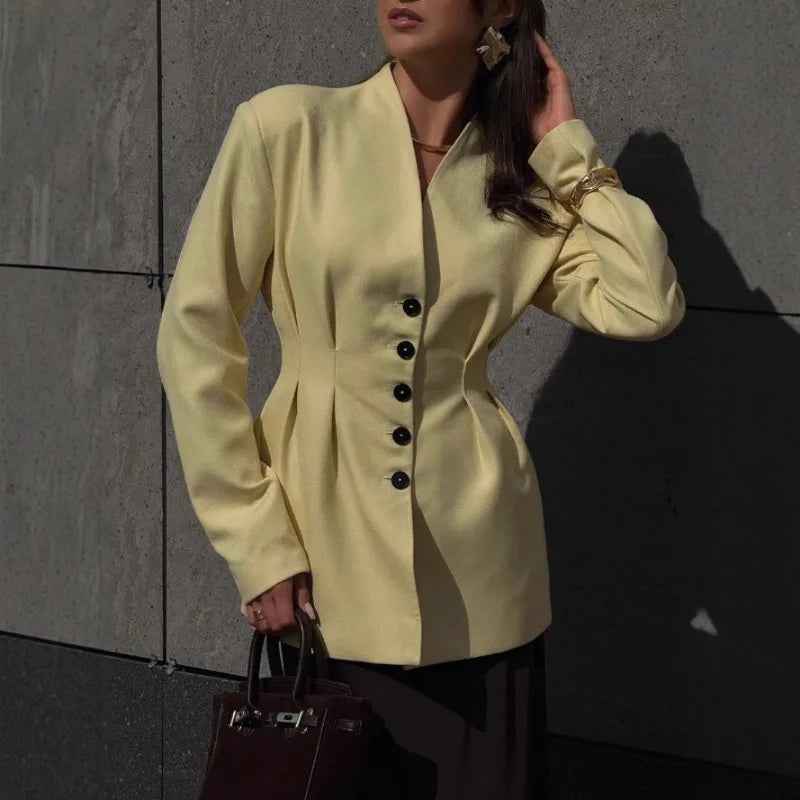 Woman wearing a light yellow blazer with black buttons against a gray wall.