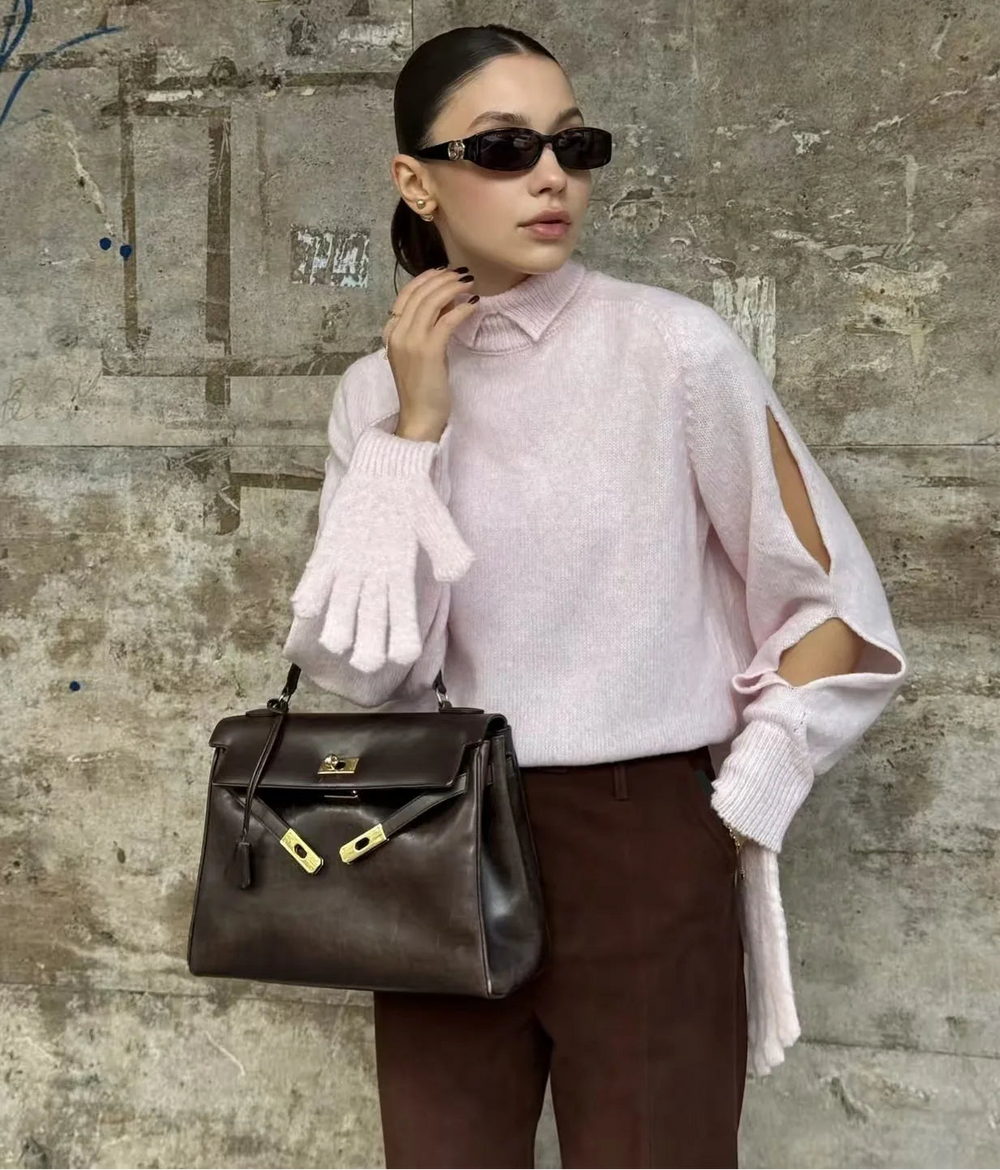 Woman wearing a light pink sweater with cut-out shoulders, brown pants, and holding a dark handbag against a textured wall.
