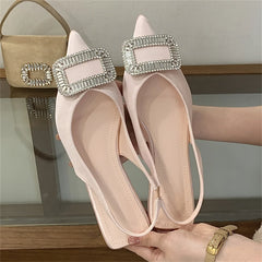 Pink pointed-toe flats with decorative buckles held by a hand on a reflective surface.
