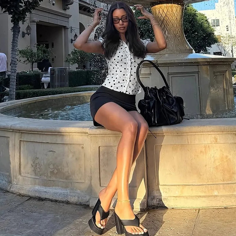 Woman sitting on a stone ledge outdoors, wearing a polka dot blouse and black skirt.