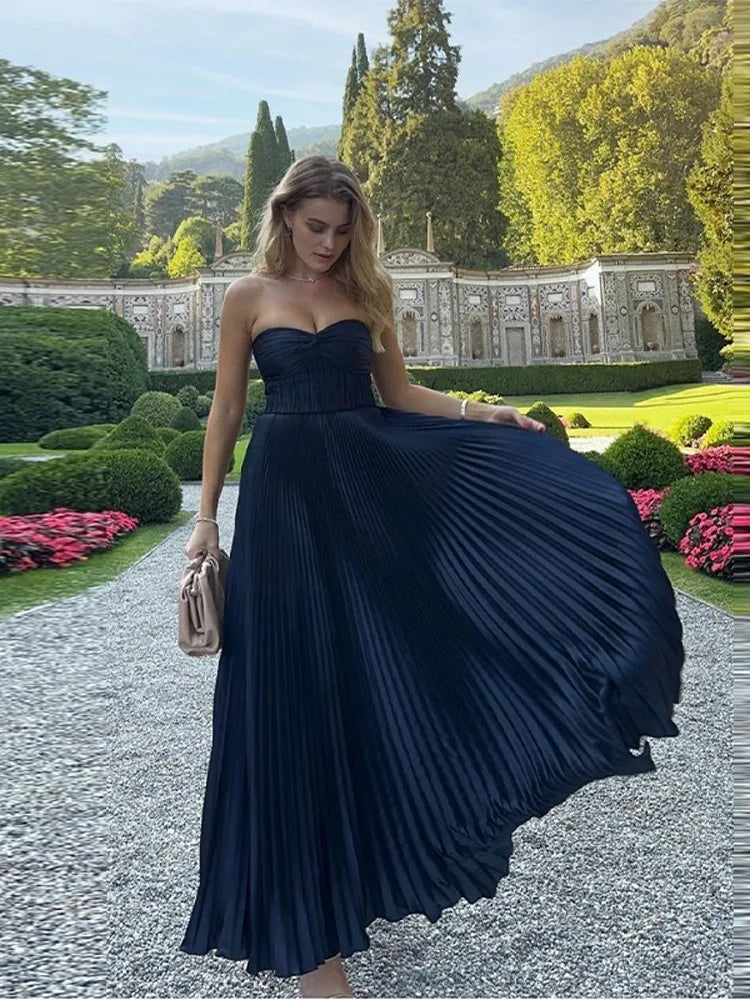 Woman in a navy pleated dress standing in a garden with greenery and a building in the background