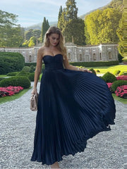 Woman in a navy pleated dress standing in a garden with greenery and a building in the background