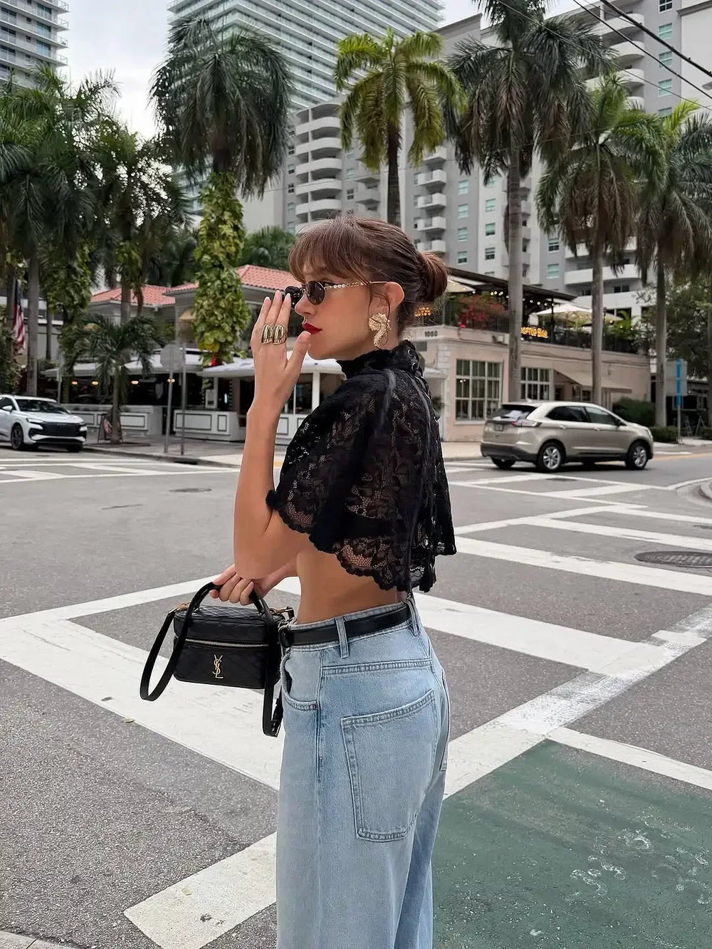 Woman in black lace top and light blue jeans standing on a city street with palm trees and buildings in the background.