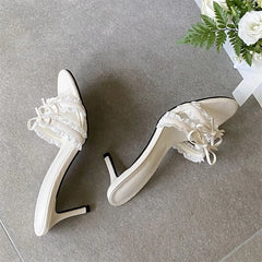 Pair of white ruffle bow stiletto heels on a grey surface.