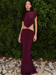 Woman wearing a burgundy evening gown with cut-out design against a green hedge background.