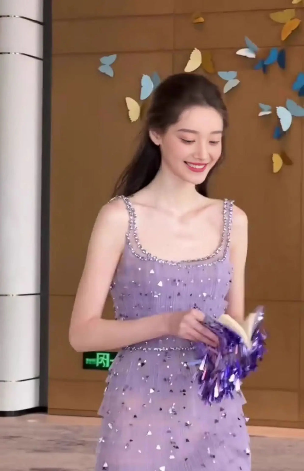 Woman in a purple sequined dress holding a book against a decorative butterfly wall.
