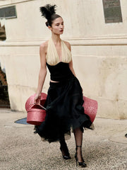 Woman in a black dress with a high ponytail holding a red suitcase against a stone wall.