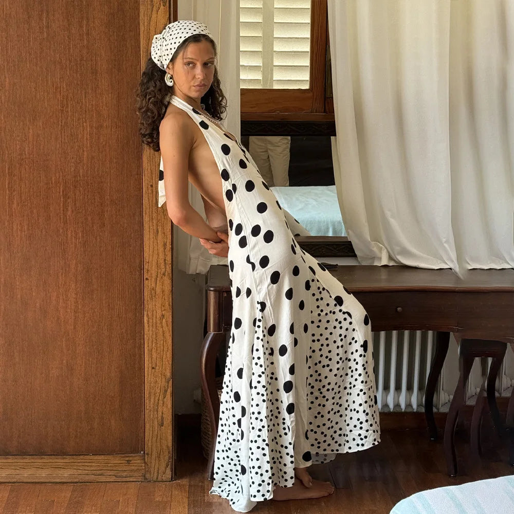 Woman in a polka dot dress standing in a room with wooden furniture and white curtains.