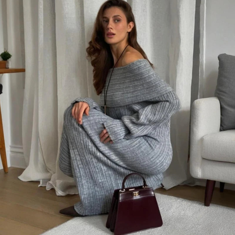 Woman in a gray sweater and pants sitting on the floor with a burgundy handbag in a room with white curtains and a gray chair.