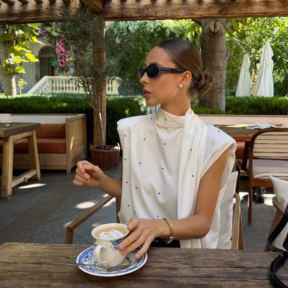 Woman sitting at an outdoor cafe table with a cup of coffee, wearing sunglasses and a white blouse.