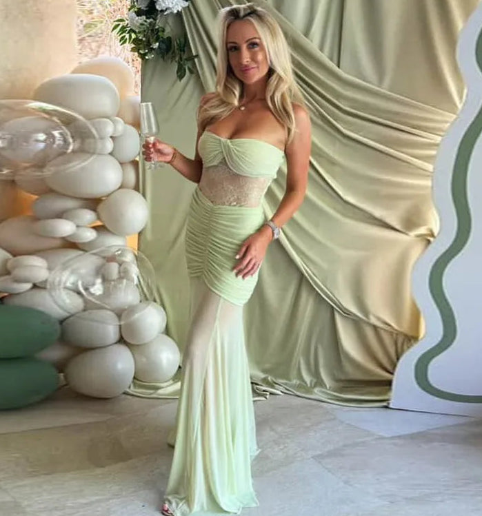 Woman in a light green dress holding a glass in front of decorative stones and draped fabric.
