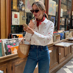 Woman in a bookstore wearing a white blouse with red patterns and blue jeans, holding a compact mirror.