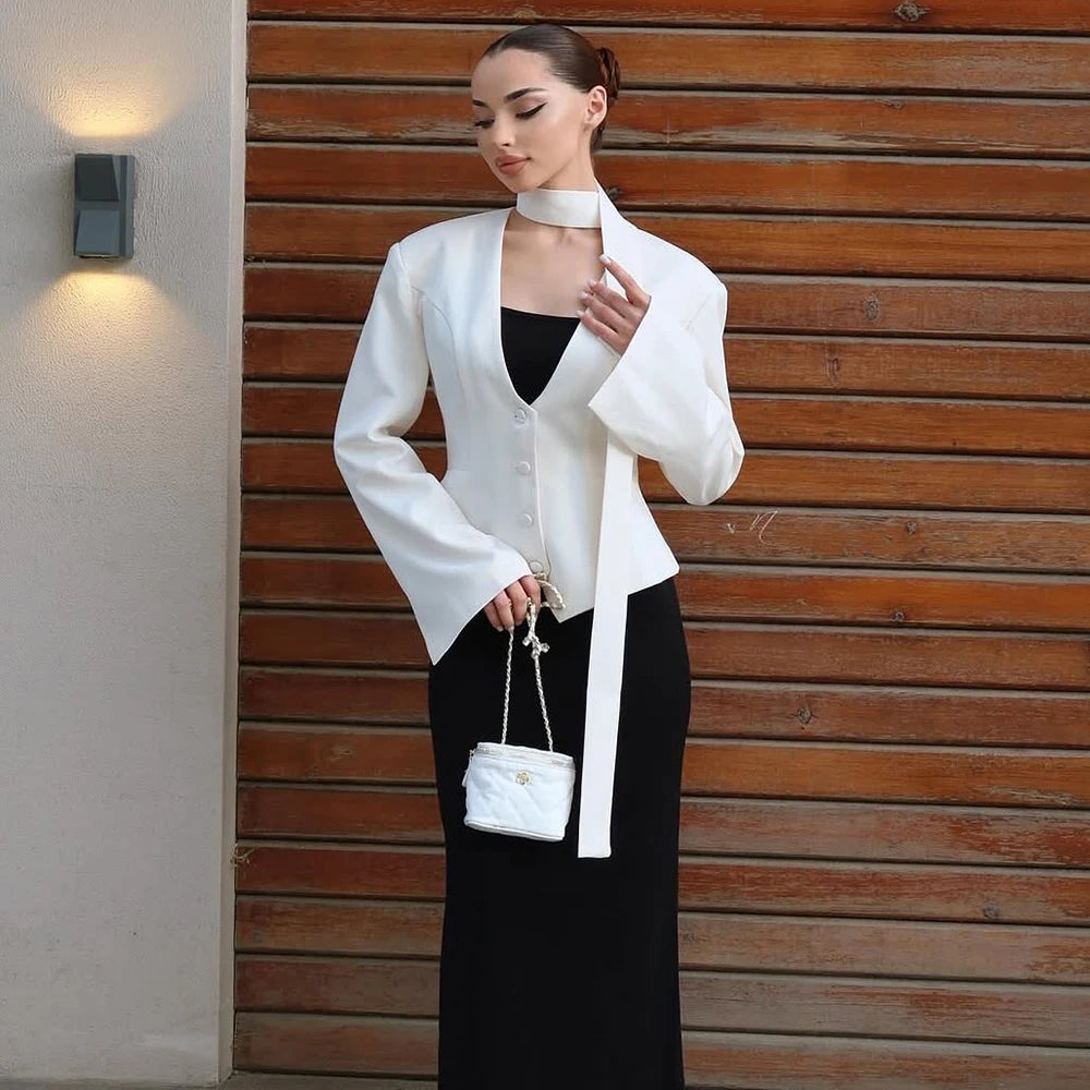Woman in a white blazer and black dress holding a white handbag against a wooden wall.