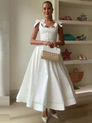 Woman in a white dress holding a beige handbag in a room with shelves.