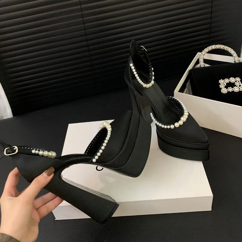 Black high-heeled shoes with pearl details held by a hand on a black surface.