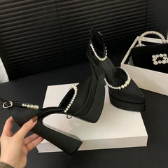 Black high-heeled shoes with pearl details held by a hand on a black surface.