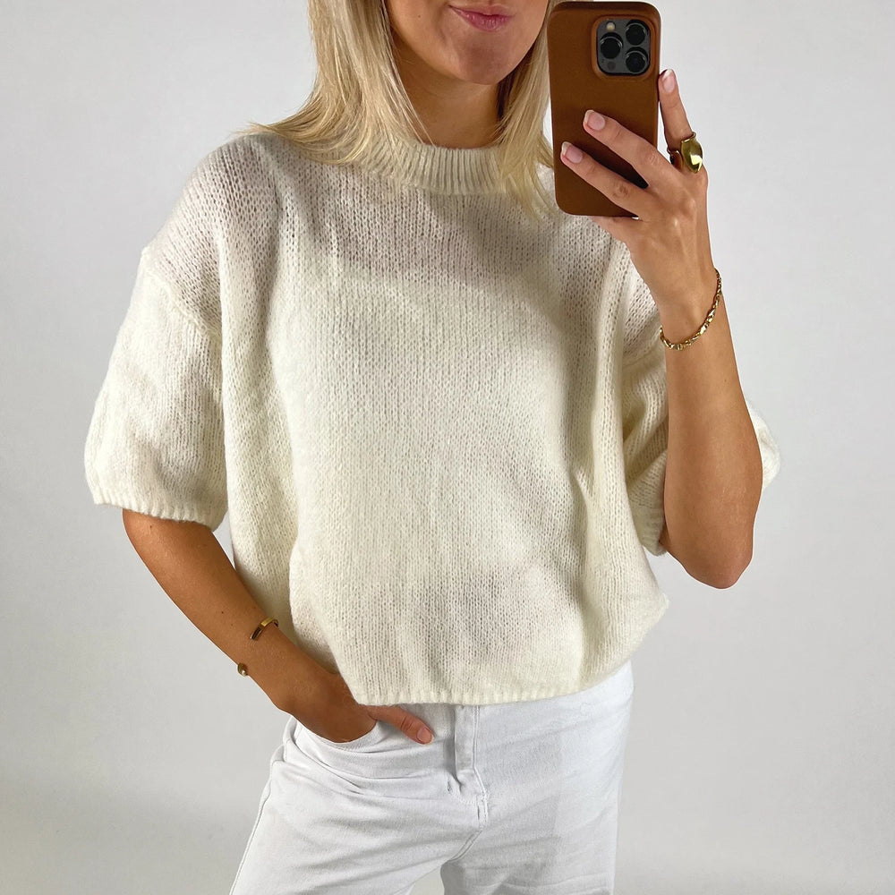 Person wearing a cream sweater and white pants, taking a mirror selfie.