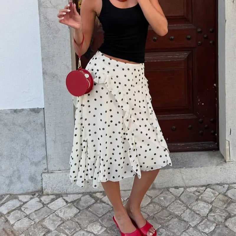 Woman wearing a black top and white polka dot skirt with a red bag, standing on a stone pavement.