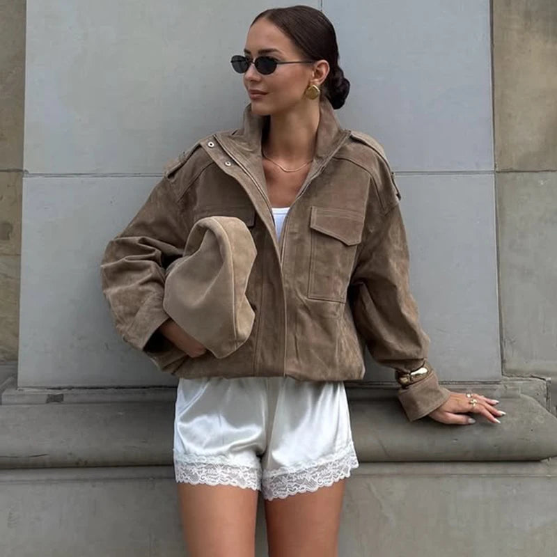 Woman wearing a brown jacket and white shorts against a gray wall.