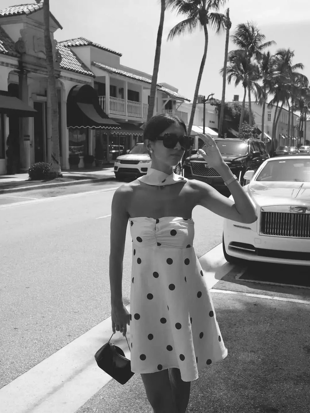 Woman in a polka dot dress taking a selfie on a street with palm trees and cars in the background.