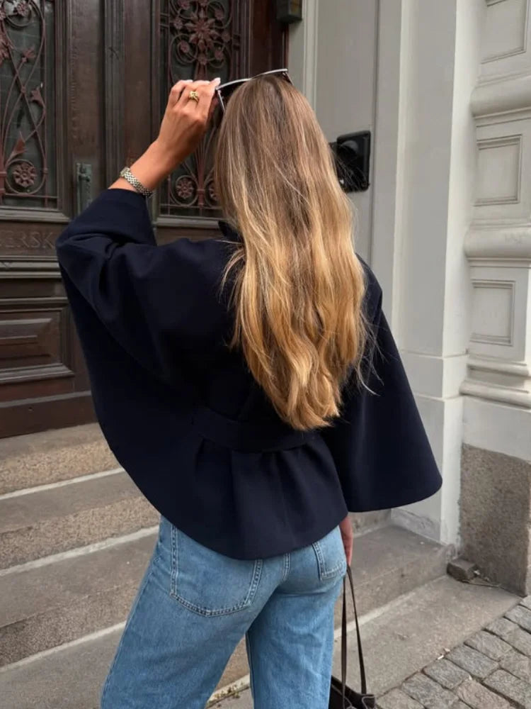 Person wearing a black jacket and blue jeans standing in front of a building entrance.