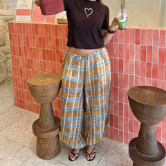 Person wearing a brown t-shirt with a heart design and plaid pants, holding a drink, standing in front of a red tiled wall.