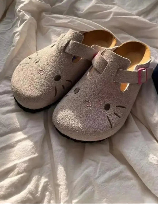 Light grey Hello Kitty flat mule slippers with pink buckle straps on a white textured surface.