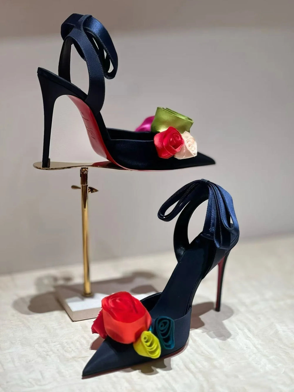 A pair of dark blue stiletto heels with colorful fabric roses and ankle straps displayed on a gold stand.