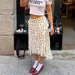 Person wearing a white crop top with text and a polka dot skirt standing on a street.