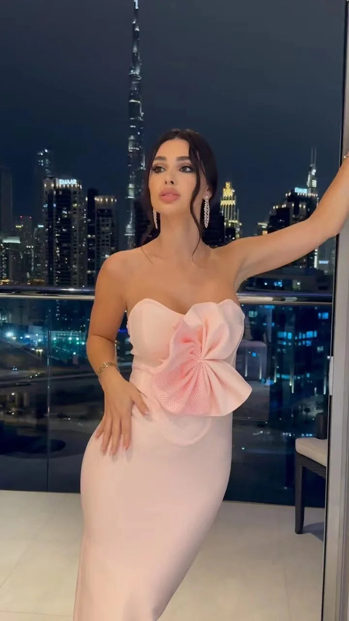 Woman in a pink dress with a large flower detail against a city skyline at night.
