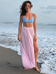 Woman in a blue top and pink skirt standing on a beach with ocean waves.