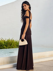 Woman in a long brown dress holding a beige clutch against a light-colored wall.