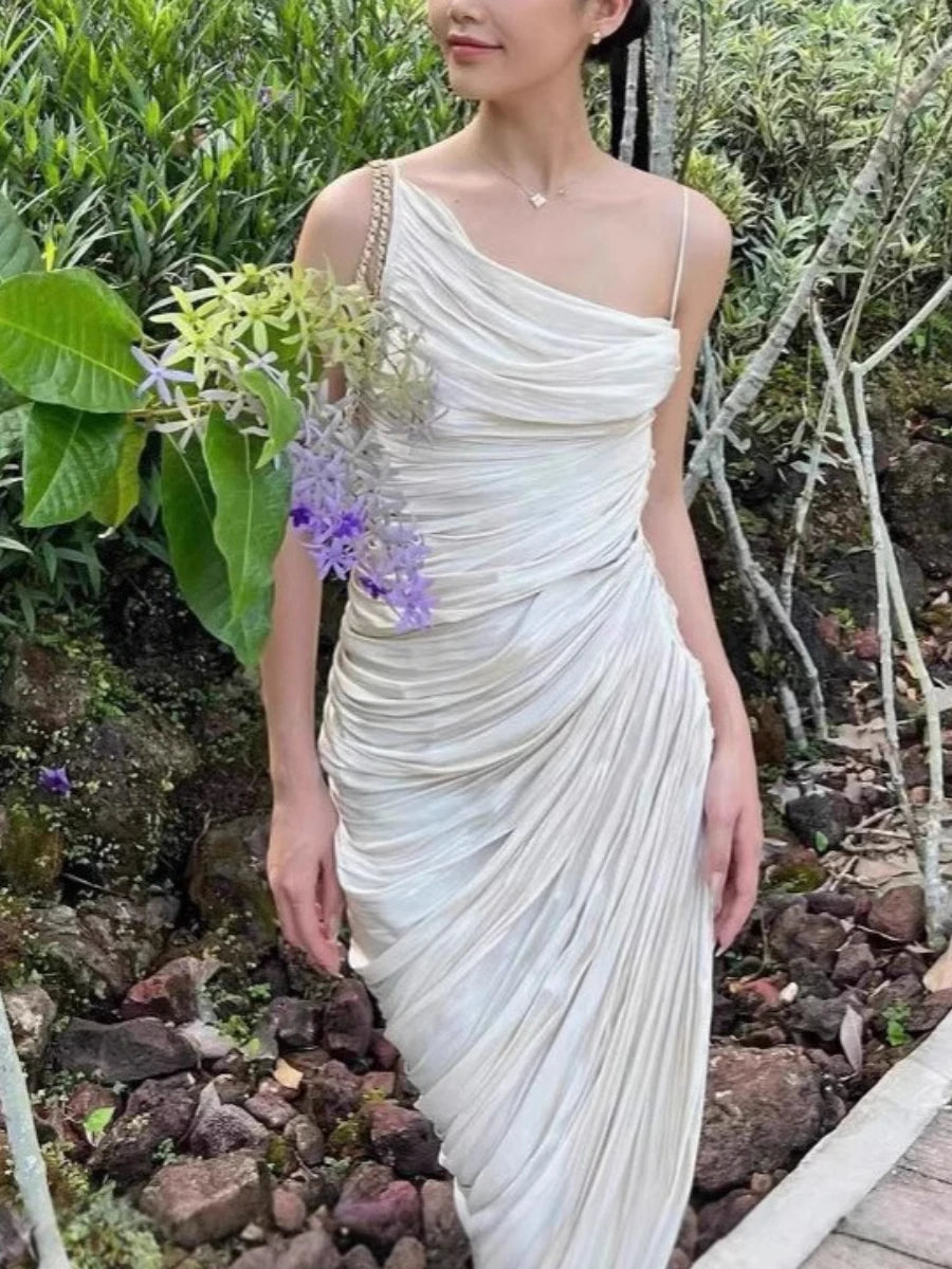 Woman in a beige one-shoulder dress standing in a natural setting with greenery.