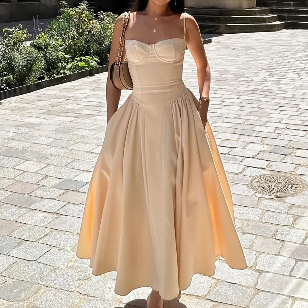 Woman wearing a beige dress standing on a stone pavement with greenery in the background