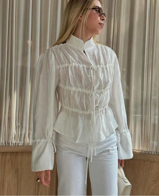 Woman wearing a white blouse with ruffled details and white pants, standing indoors.