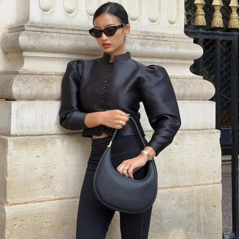 Woman in black outfit holding a matching handbag against a stone wall.