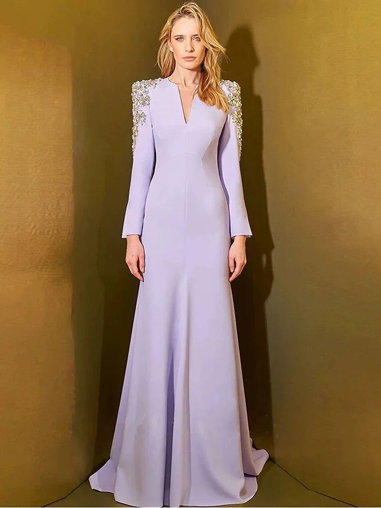 Woman wearing a long, light purple evening gown with embellished shoulders against a gold background
