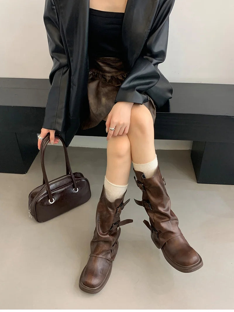 Person wearing brown knee-high boots and holding a brown handbag on a plain background