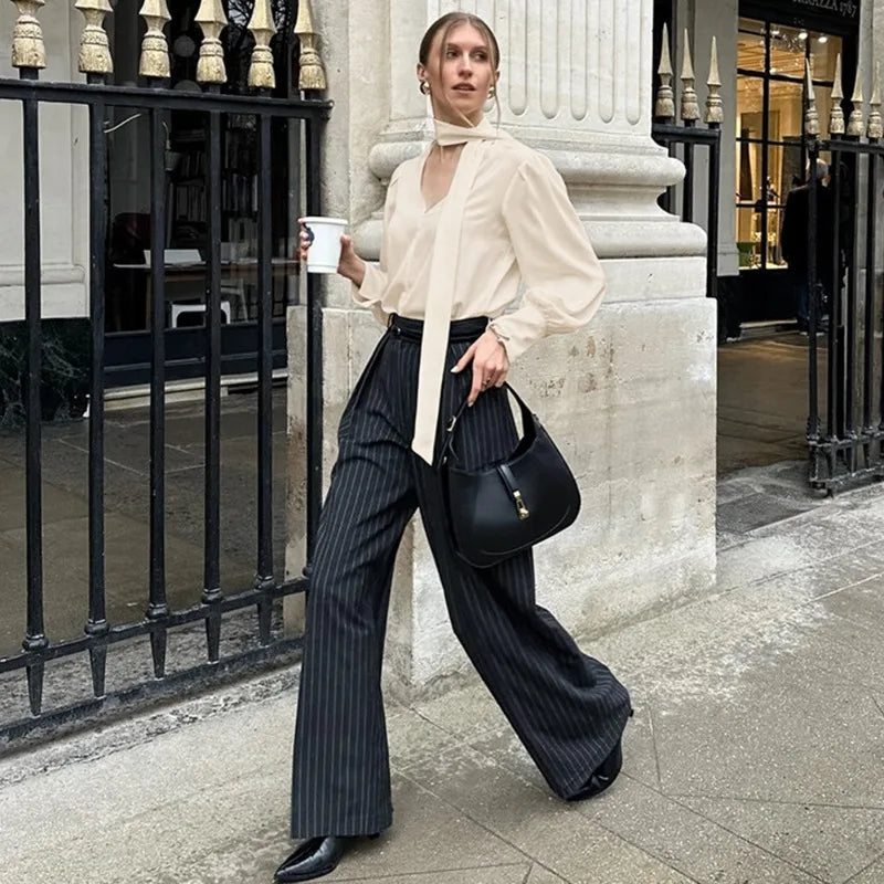 Woman in a stylish outfit with a white blouse and black pinstripe pants, holding a black handbag and a cup, standing against a city street background.