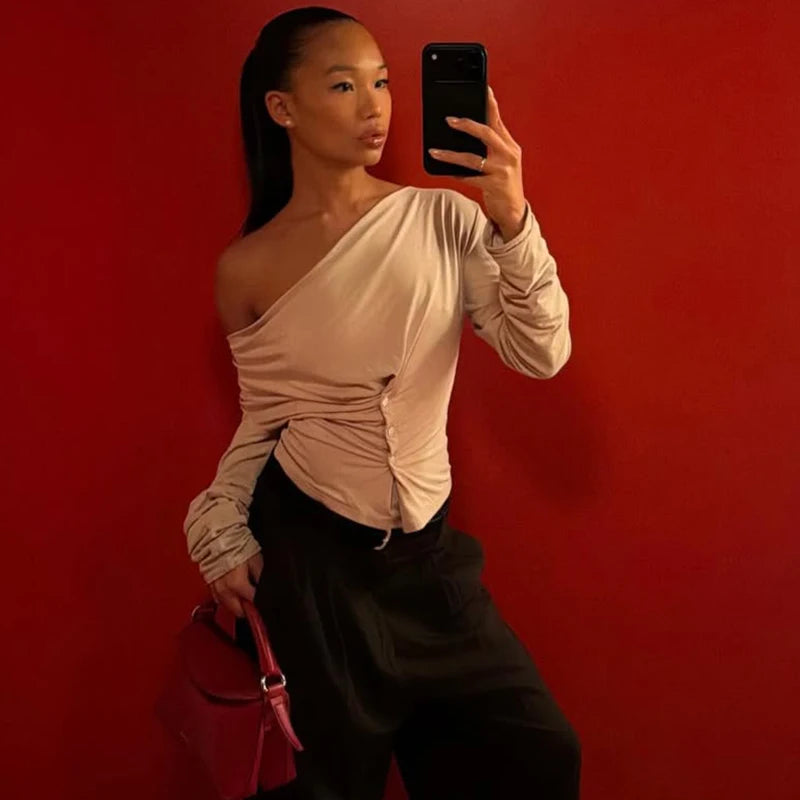 Woman taking a mirror selfie against a red wall, wearing a beige one-shoulder top and black pants.