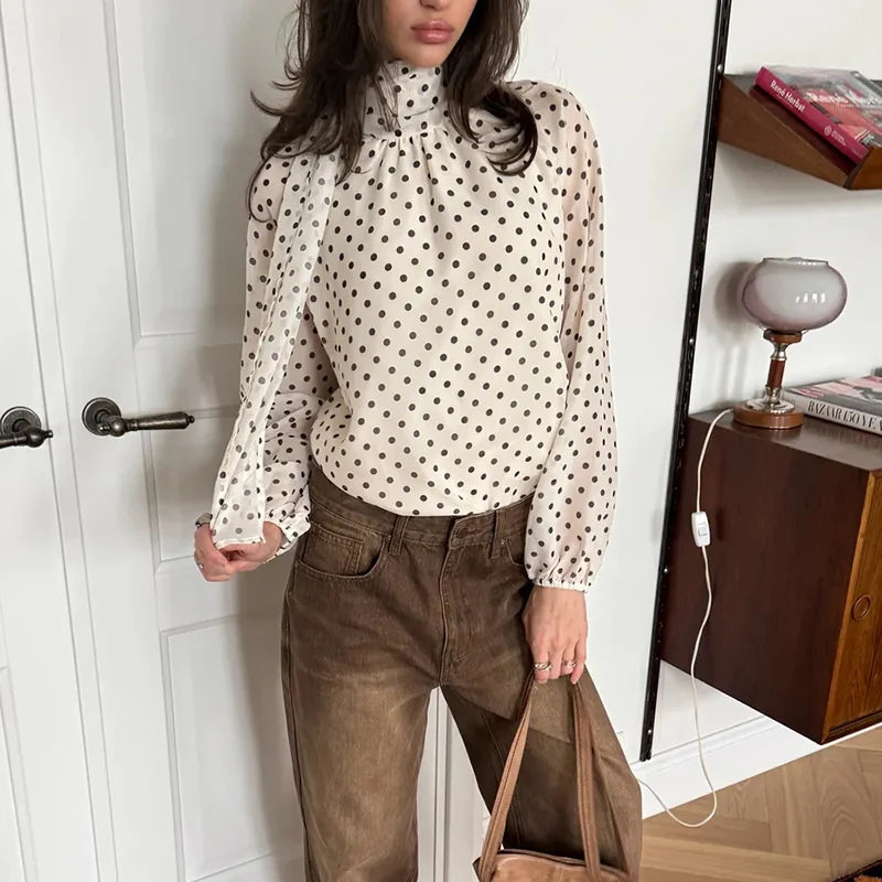 Woman wearing a polka dot blouse and brown pants in a room with a shelf and door.