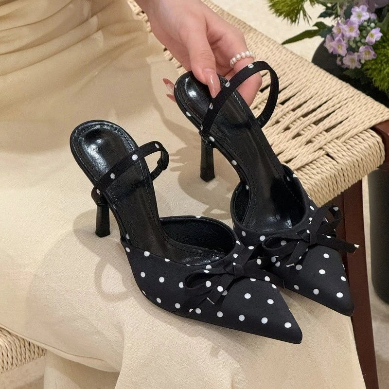 Black high-heeled shoes with white polka dots and bows held by a hand on a beige surface.
