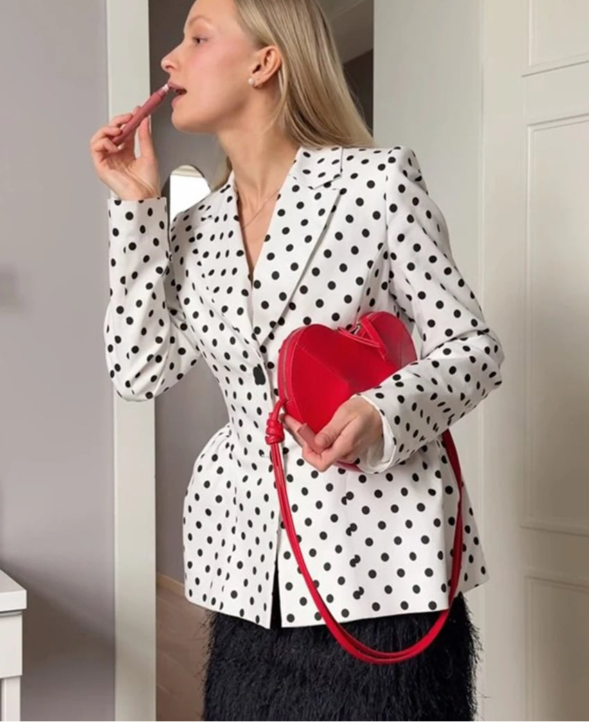 Woman in polka dot blazer with red handbag applying lipstick indoors