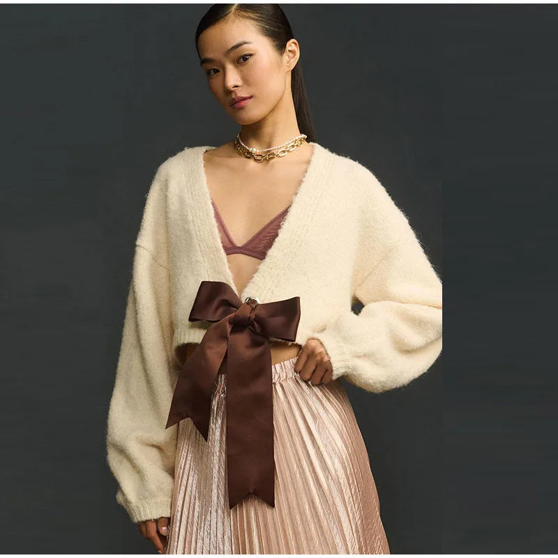 Woman wearing a cream cardigan with a brown bow and pleated skirt against a dark background